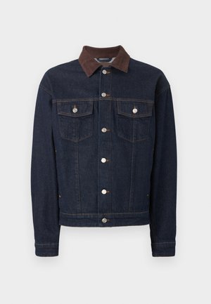 Jeune homme portant une veste en jean foncé sur une chemise blanche à boutons, un pantalon beige à jambes larges et des chaussures en cuir noires, debout les mains dans les poches.