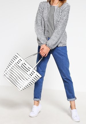 Osoba ubrana w szarą fakturowaną kurtkę, koszulę w paski, podwinięte niebieskie dżinsy, białe trampki, trzymająca dużą białą perforowaną torbę na ramię.