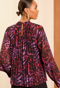 Blusa trasparente nera con una vivace stampa animale in viola e rosso. Presenta delle pieghe sul retro e un collo rotondo con apertura a chiave. Maniche lunghe.