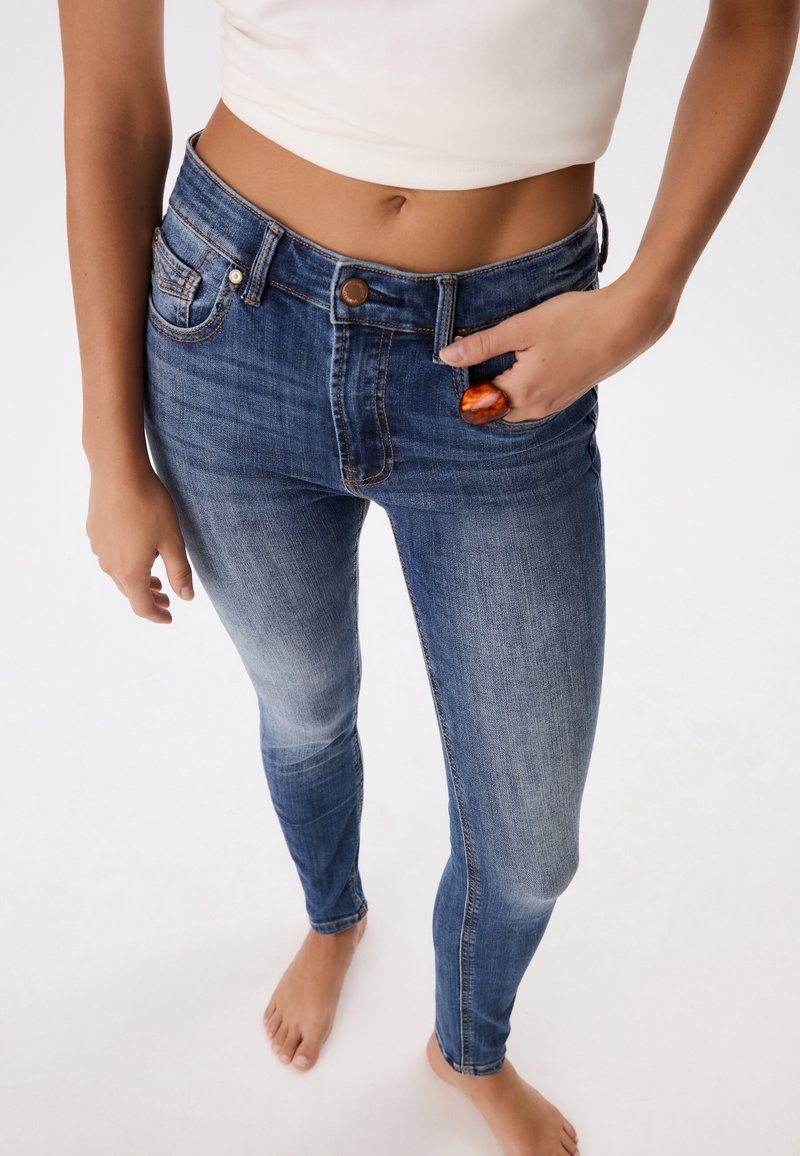Faded blue denim skinny jeans with a high waist, five pockets, and a metal button closure. Model holds a small orange stone.