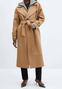 Płaszcz z wełny wielbłądziej z paskiem w talii, dużymi klapami i szarym, odpinanym kapturem. Zawiera dwie boczne kieszenie oraz mankiety zapinane na guziki.