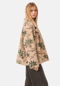 Chaqueta de forro polar beige con patrones de hojas verdes y acentos de lentejuelas, con un corte holgado y una textura suave. Los bordes son redondeados.