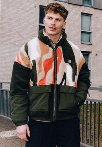Chaqueta de forro polar con patrón abstracto en tonos de naranja, blanco y verde; cierre frontal con cremallera; dos grandes bolsillos frontales; tejido texturizado.