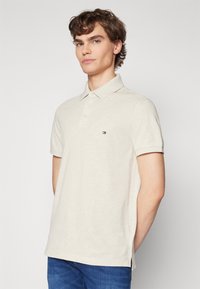 Polo beige realizzato in tessuto morbido con collo a polo, tre bottoni e un piccolo logo sulla parte sinistra del petto. Maniche corte e spacco sui lati.
