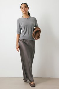 Maglione corto grigio a maniche corte abbinato a una lunga gonna di raso marrone scuro. La modella tiene una borsa in suede marrone e indossa scarpe flat marroni.