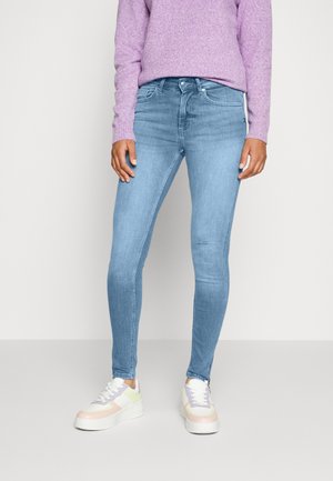 Personne portant un jean skinny bleu clair, un pull lilas et des baskets blanches avec des accents pastel, debout devant un fond clair uni.