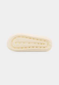 Semelle d'une chaussure beige avec un motif de grip texturé et de petits accents ronds jaunes disposés en deux lignes le long du périmètre et du centre.