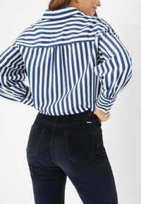 Blauw-wit gestreepte shirt met een losse pasvorm, met een rugpand en knopenmanchetten. Gecombineerd met zwarte hooggerimpelde broek.