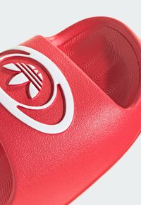 Raudona šlepetė su tekstūruotu viršumi, ant kurio yra ryškus baltas logotipas. Lygus paviršius su bangelių rašto padu, suteikiančiu sukibimą.