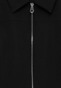 Veste noire avec une fermeture éclair métallique argentée s’étendant verticalement du col jusqu’au milieu, avec un col de chemise standard.