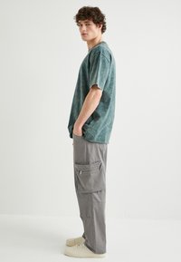 Maglietta oversize tie-dye verde a maniche corte, abbinata a pantaloni cargo grigi chiari con cerniere e tasche, completata da sneakers beige.