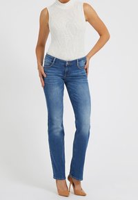 Jeans en denim bleu avec une coupe slim, un léger effet délavé et une taille mi-haute, associés à un haut sans manches texturé blanc et des talons beiges.