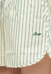 Gros plan sur un tissu crème avec de fines rayures verticales vertes et un texte brodé vert près du bord arrondi de l'ourlet.