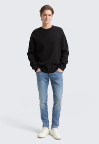 Sudadera negra de cuello redondo, combinada con jeans azules y zapatillas blancas. La tela parece suave, con un corte relajado y líneas limpias.
