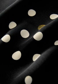Schwarzer Stoff mit weißen Punkten in verschiedenen Größen; weiche Textur zeigt Falten und einen subtilen Glanz; ein Punkt ist golden.