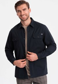 Camisa de botones azul marino en una mezcla de algodón con dos bolsillos en el pecho, mangas largas y un sutil acento de herrajes. Lleva puesta una camiseta beige por debajo.