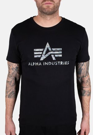 Man wearing black t-shirt with faded Alpha Industries logo and text, showing tattooed arms against plain background.