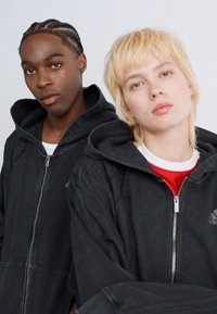 Two people wearing black hooded jackets, standing close; one with braided hair, the other with short blonde hair and a red shirt underneath.