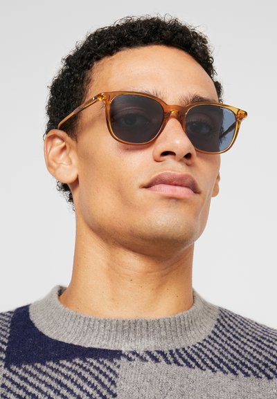 Ochelari de soare tip cat-eye cu nuanță maronie și lentile închise, având o ramă din chihlimbar translucent. Designul este modern, cu linii clare și hardware minim.