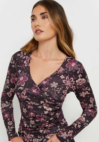 Pitkähihaista tumman violetista paita, jossa on kukkakuviota. Pehmeä kangas, jossa v-pääntie ja kokoontumisdetaileja vyötäröllä, korostaen istuvaa muotoa.