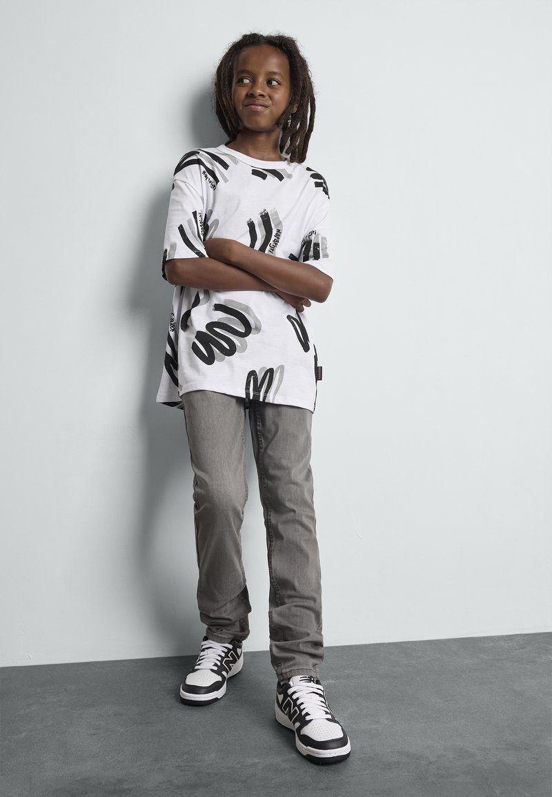 Camiseta blanca con patrones abstractos en negro, combinada con jeans grises y zapatillas en blanco y negro. Diseño simple con corte holgado y mangas cortas.