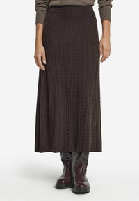 Kvinna som bär en lång mörkbrun kabelstickad kjol tillsammans med glänsande mörkburgundy stövlar som når till mitten av vaden, stående mot en vit bakgrund.