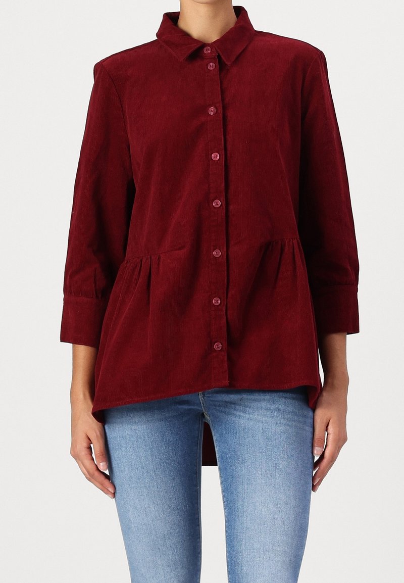 Femme portant une chemise en velours côtelé bordeaux à manches longues et à boutons avec col, associée à un jean bleu clair, posant devant un fond uni.