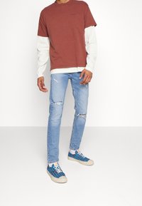 Vinröd T-shirt med korta ärmar över en vit långärmad tröja, kombinerad med ljusblå slitna jeans och färgglada mönstrade sneakers.