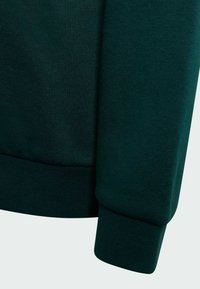 Puño de sudadera verde oscuro, textura acanalada y acabado de tela suave. El puño es un poco más ancho, lo que añade suavidad al diseño general.