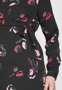 Svart klänning i tyg med blommönster i röd, rosa och vit. Inkluderar ett knytbälte i midjan och långa ärmar med knappribbar.