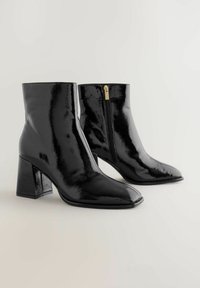 Black ankle boots made of patent leather, featuring a square toe, block heel, and side zipper with gold hardware detail.