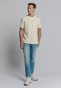Gestreiftes Kurzarm-T-Shirt in sanftem Beige und Creme, kombiniert mit hellblauen Jeans und weißen Sneakers, vor einem grauen Hintergrund stehend.