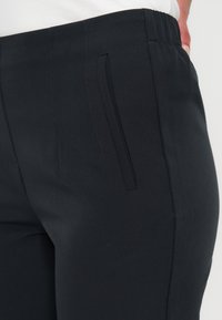 Personne portant un pantalon noir ajusté avec une ceinture élastique, une poche fendue sur le côté et une chemise blanche rentrée, vue des hanches vers le bas.
