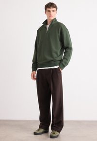 Felpa verde con zip e colletto alto, abbinata a pantaloni marroni larghi e sneakers verdi, su uno sfondo neutro.