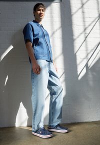 Camicia in denim a maniche corte blu, vestibilità rilassata. Jeans leggeri blu, gamba dritta. Zoccoli blu navy con righe rosse e bianche. Texture liscia, stile casual.