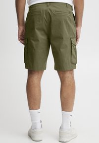 Shorts cargo verts olive en tissu tissé, avec des poches latérales, une coupe droite et une taille confortable. Portés avec des chaussettes blanches et des baskets.