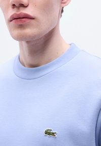 Φωτεινή γαλάζια φούτερ με στρογγυλή λαιμόκοψη και ριμπ μανσέτες. Διαθέτει μικρό κεντητό λογότυπο κροκόδειλου στο στήθος. Λεία υφή.