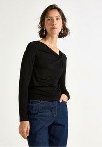 Svart långärmad topp med snodd halsringning, mjuk textur och figurnära design, ihop med vida blå jeans.