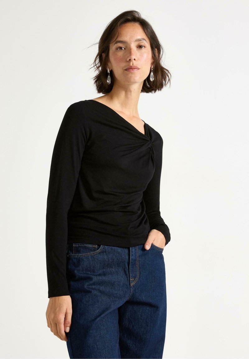 Svart långärmad topp med snodd halsringning, mjuk textur och figurnära design, ihop med vida blå jeans.