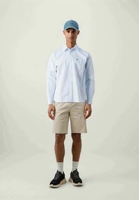 Camisa de manga longa azul clara com riscas, calções bege, sapatilhas pretas e um boné azul, numa pessoa de pé contra um fundo neutro.