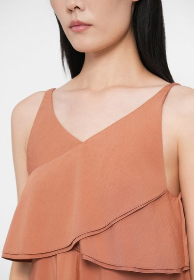 Γυναίκα που φοράει αμάνικο τοπ σε καφέ-πορτοκαλί χρώμα με εναλλασσόμενες λεπτομέρειες σε ρίγες και V-λαιμόκοψη, φαίνεται από τους ώμους μέχρι το πηγούνι.