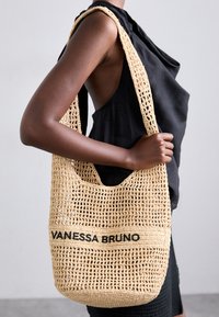 Geantă de toată ziua din fână țesută, bej, cu un model de țesătură deschisă. Prezintă un logo negru "VANESSA BRUNO" pe o bandă orizontală în apropierea centrului.