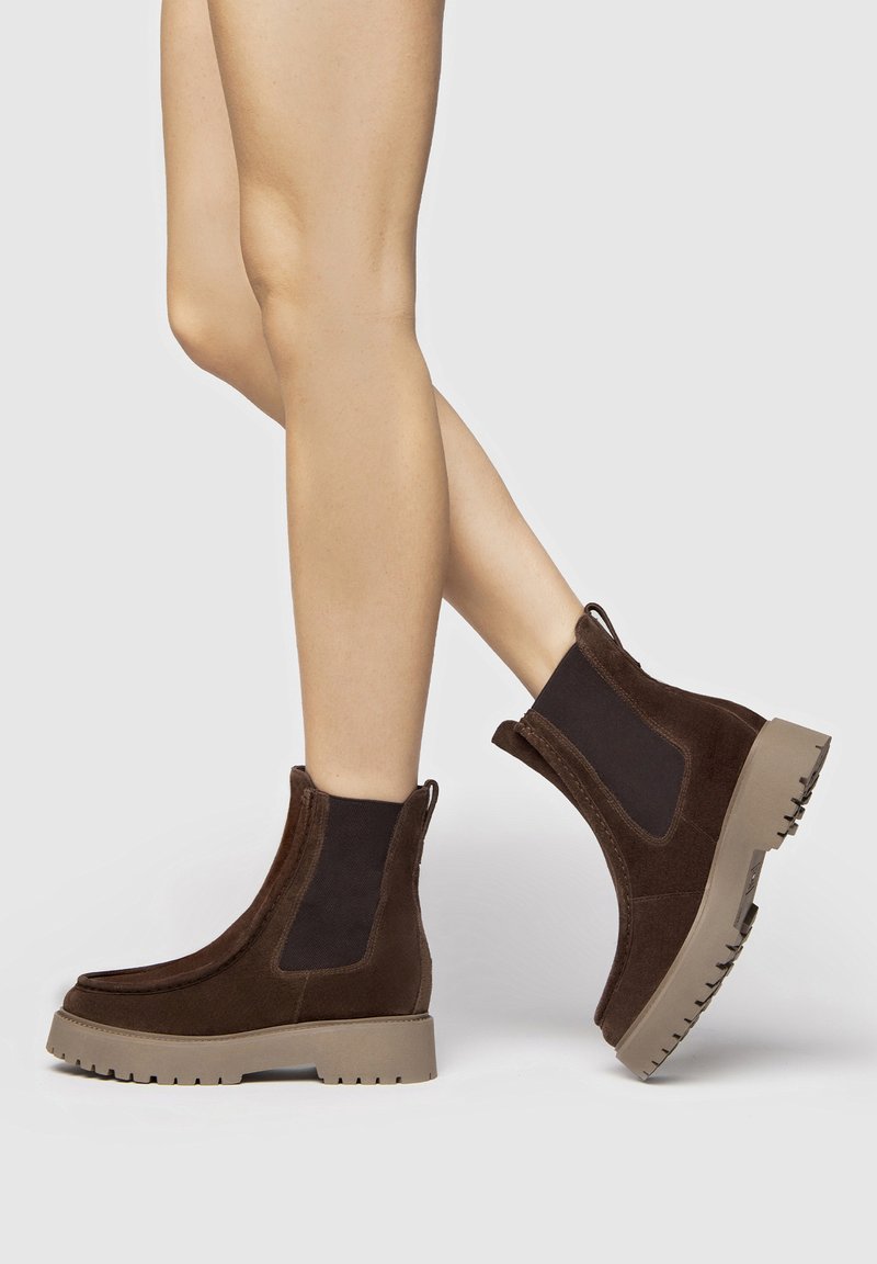 Bottines en suède marron avec des panneaux élastiques, semelles en caoutchouc beige chunky et accents texturés. Design unique pour le style et le confort.