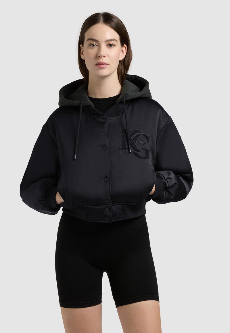 Femme portant une veste noire courte à capuche avec boutons et un grand logo KG, associée à des shorts de cyclisme noirs, debout devant un fond uni.