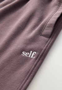 Fioletowe dresy wykonane z miękkiego materiału, z boczną kieszenią i białym haftowanym logo "self." przy dołku.