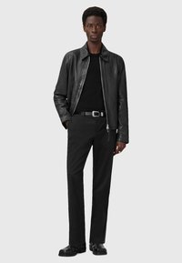Schwarze Lederjacke mit Kragen, getragen über einem schwarzen Pullover. Schwarze Hose und schwarze Schuhe komplettieren das Outfit und betonen einen monochromen Look.