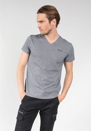 Homme portant un t-shirt gris à col en V avec le logo "DEELUXE" et un pantalon cargo noir, debout les mains dans les poches en regardant sur le côté.