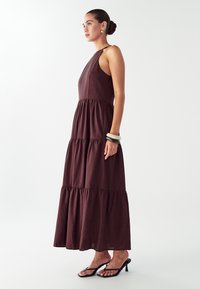 Nainen seisoo kyljelleen yllään hihaton kerroksellinen tummanruskea maksimekko, mustat korkokengät, korvarenkaat ja päällekkäin asetellut rannekorut yhdessä ranteessaan.