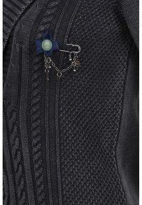Tela de punto negra con patrones texturizados. Un broche de cinta azul oscura con un centro verde y cadenas de plata colgantes añade detalle.