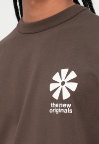 Ruda medvilnė marškinėliai su baltu gėlių logotipu ir tekstu "nauji originalai" atspausdintu kairėje pusėje. Su apvalia apykakle.